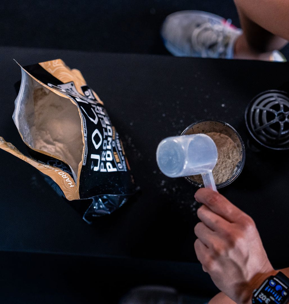 Overhead: a scoop of protein powder mid-pour into the shaker, open bag beside it