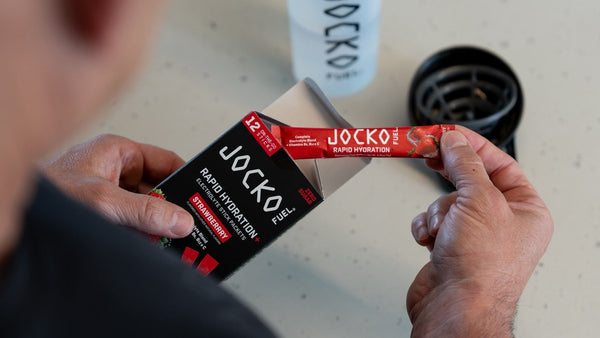 Hands pulling a Strawberry Rapid Hydration+ electrolyte stick from the 12-count box beside a Jocko Fuel shaker