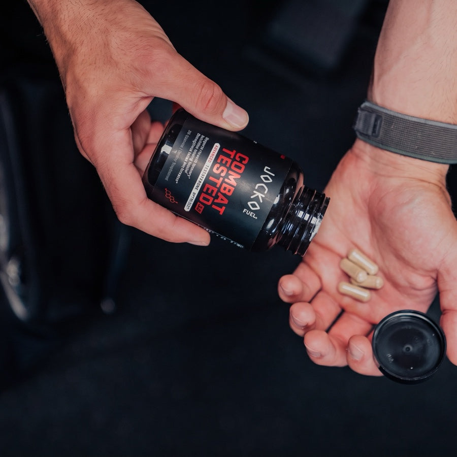 Four capsules poured from a Combat Tested 3.0 bottle into an open palm, gym bag behind