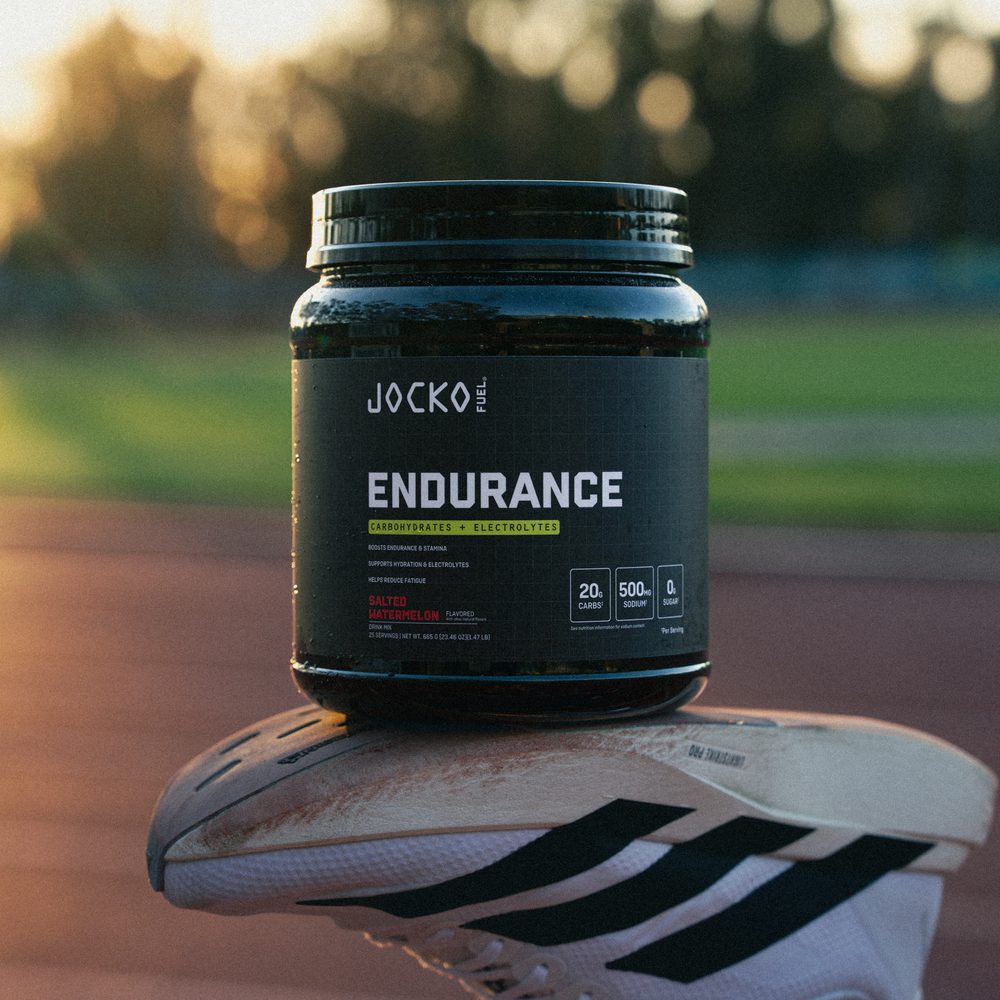 A tub of Jocko Fuel Endurance balanced on the sole of a raised running shoe, with a track and sunlit trees behind it.
