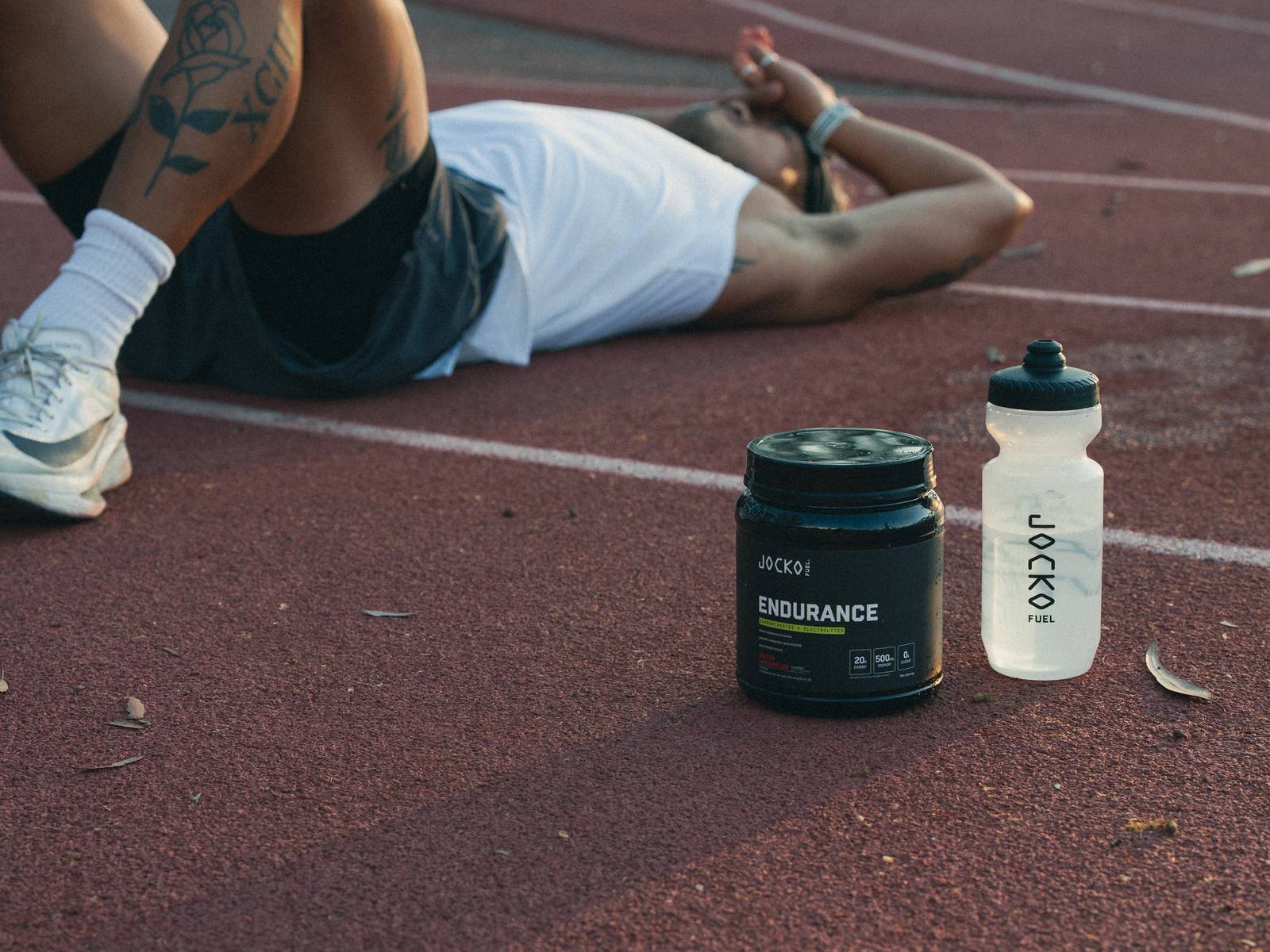 A runner lying flat on his back on a running track between efforts, with a tub of Endurance and a shaker bottle standing on the track beside him.