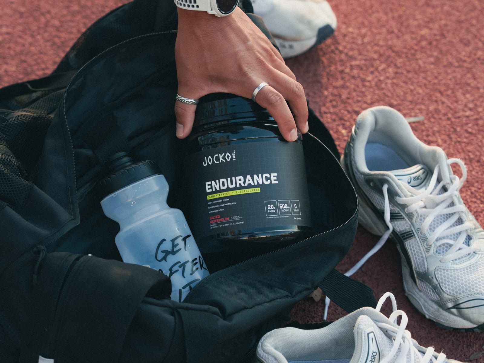 A hand lifting a tub of Jocko Fuel Endurance out of an open gym bag on a running track, next to a shaker bottle and a pair of running shoes.