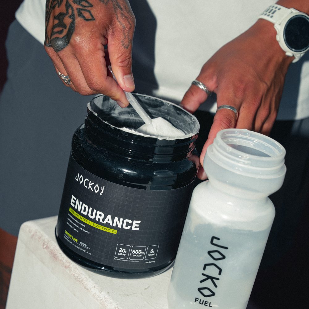 A scoop of Endurance powder being lifted from the open tub on a bench, with an uncapped shaker bottle held ready beside it.