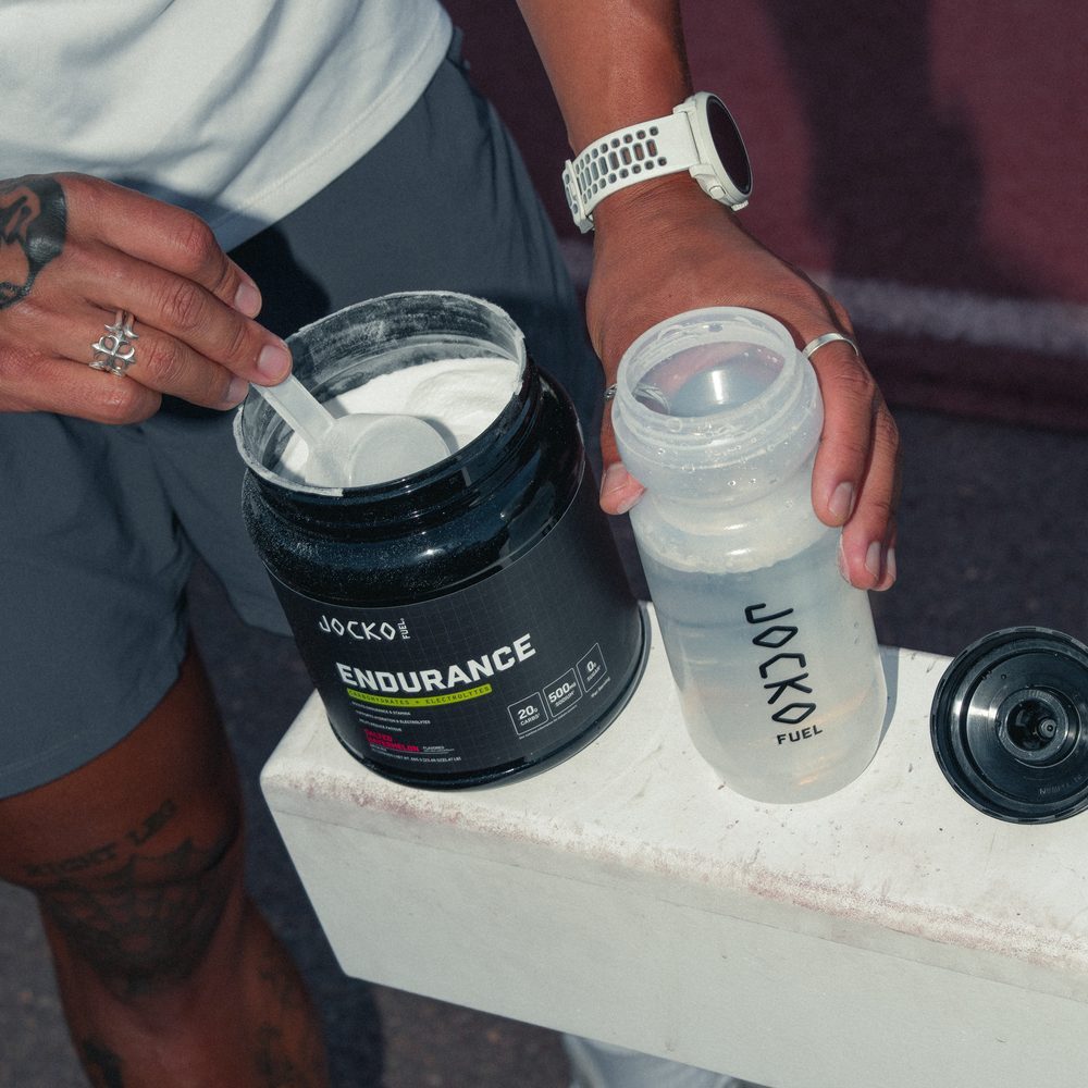 One hand scooping from the open Endurance tub while the other holds the shaker bottle with its cap off, ready to mix.