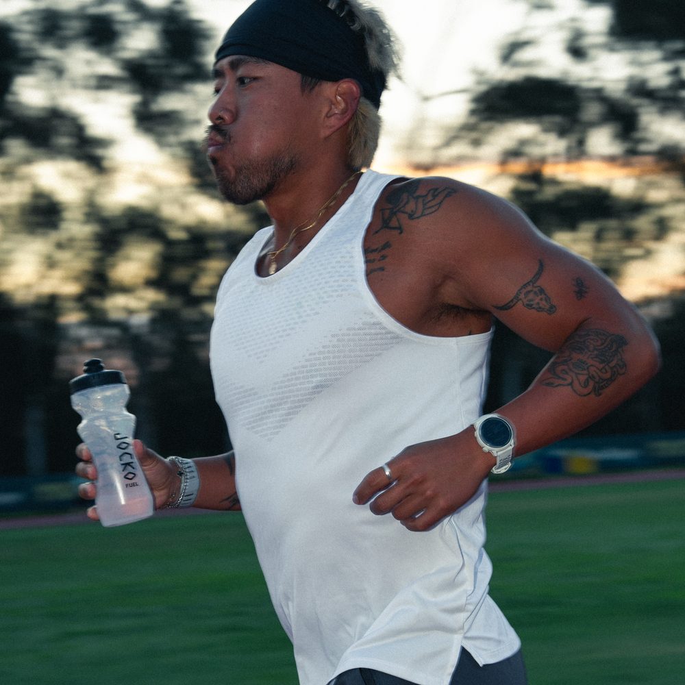 A runner mid-stride on a track at dusk, cheeks puffed on an exhale, carrying a Jocko Fuel shaker bottle in one hand.