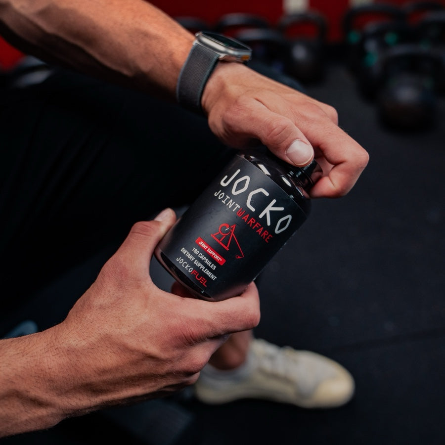 A man seated in a gym opening a bottle of Jocko Joint Warfare, kettlebells behind him