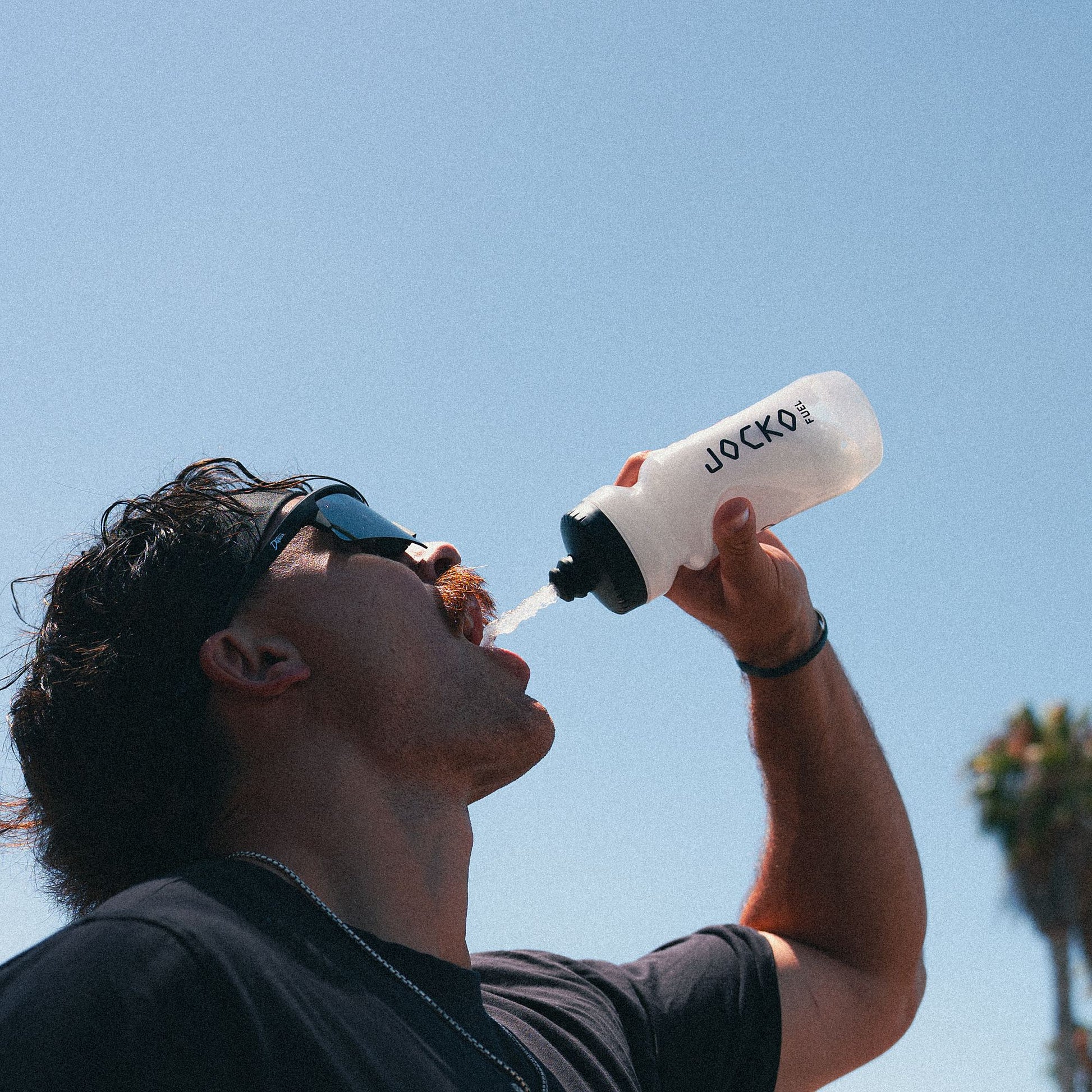 Person drinking from a Jocko water bottle against a clear blue sky