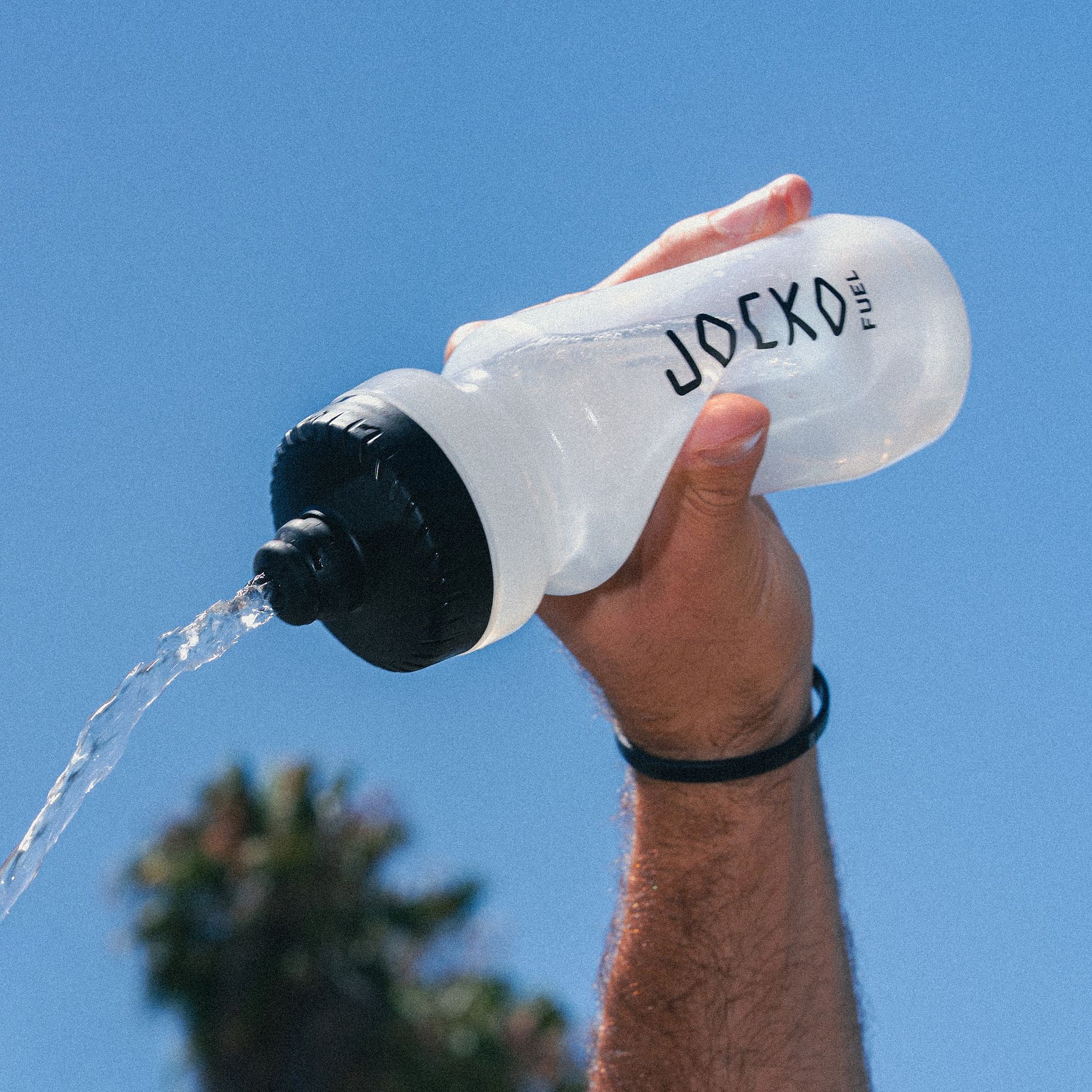 Hand holding a JOCKO Fuel water bottle with water flowing against a clear blue sky.