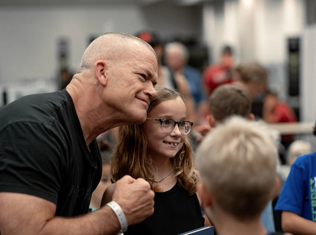 Jocko Willink cheek to cheek with a smiling young Warrior Kid, other kids gathered around