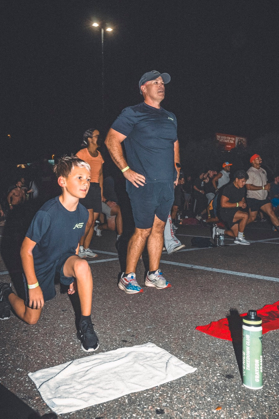 A boy and his dad doing lunges together at a night workout in a parking lot