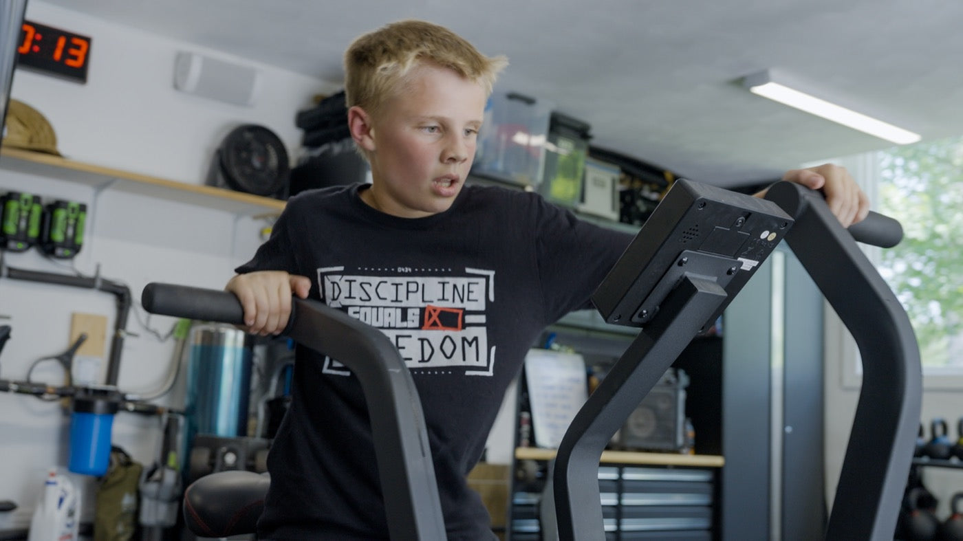 A boy in a Discipline Equals Freedom shirt working on an air bike in a garage gym